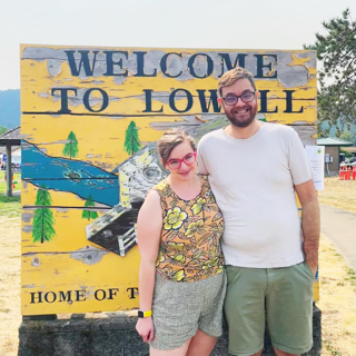 A tall man and a shorter woman pose in front of a sign for Lowell
