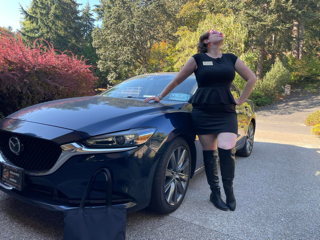 A woman in a black dress and boots leans on a car