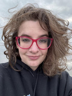 A woman with messy hair and pink glasses looks into the camera